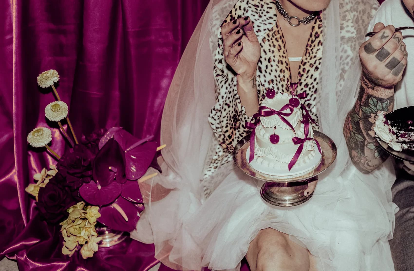A bride in a veil and leopard-print coat holding a small wedding cake tied with magenta ribbons